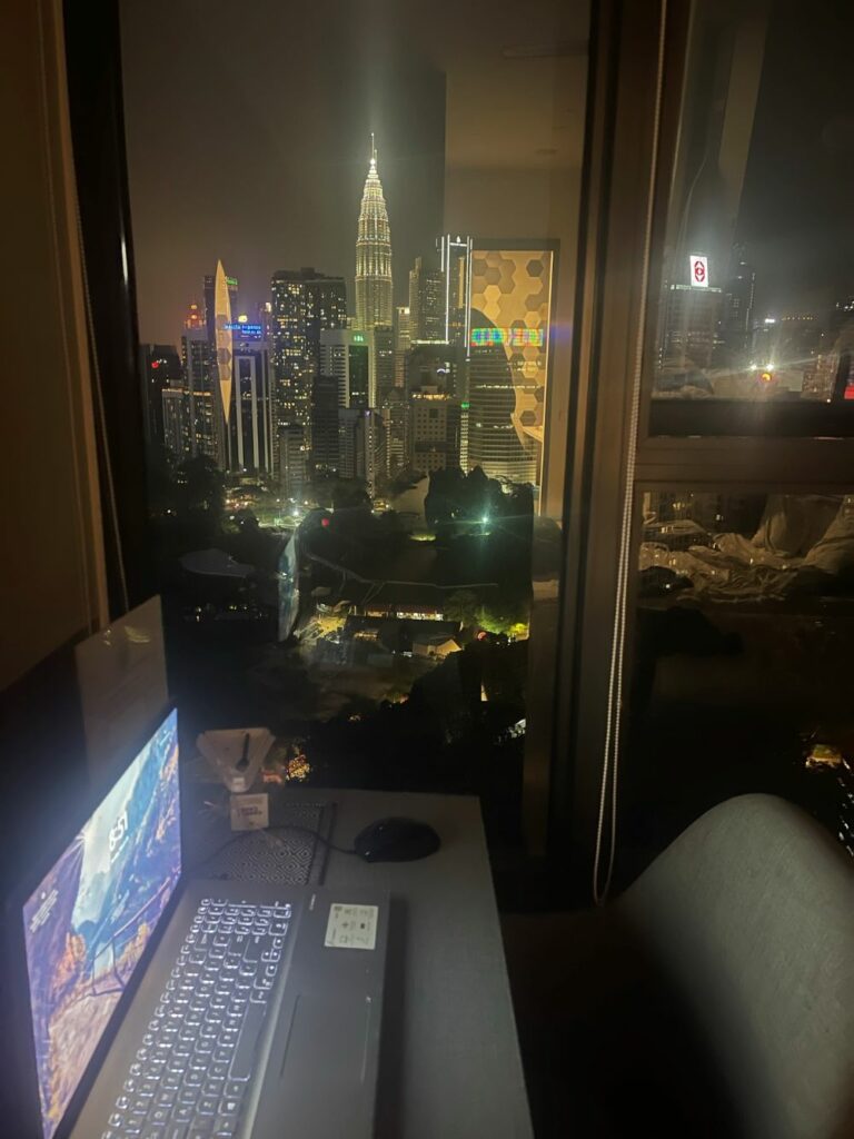 A laptop on a desk in front of a window in a hotel in Kuala Lumpur. The Petronas Twin Towers are visible outside at night.