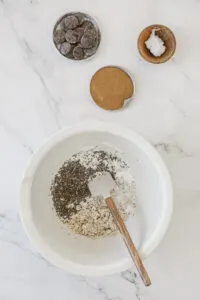 A mixing bowl with oats and almond milk and chia seeds being mixed together. Peanut butter and chocolate chips in bowls next to it.