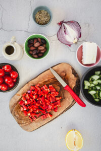 Overhead shot of the ingredients for a Greek pasta salad with diced red pepper on a chopping board