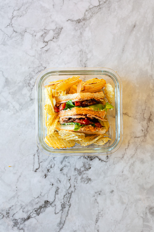 club sandwich in a storage container surrounded by chips