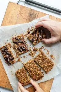 Granola Bars With Melted Chocolate drizzled on them are shown on top of a wooden chopping board