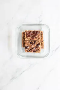 Homemade granola bars drizzled with melted chocolate are shown in side a glass storage container